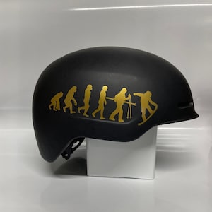 Op de afbeelding: Een zwarte skihelm met een gouden sticker van de evolutie van de mens, met aan het einde van de rij een skiër.