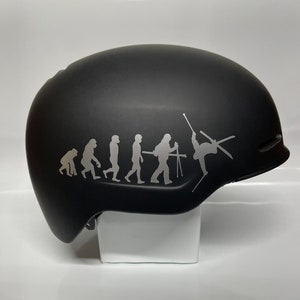 May include: A black ski helmet with a silver decal of the evolution of man, ending with a skier doing a trick.