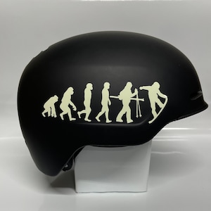 May include: A black snowboard helmet with a light green evolution graphic. The graphic depicts the evolution of man, culminating in a snowboarder. The helmet is designed for winter sports and provides head protection.
