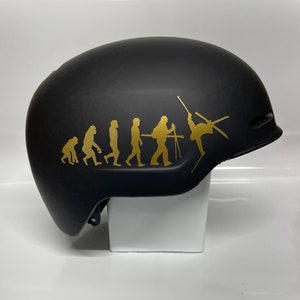 Puede incluir: Casco de esquí negro con un gráfico dorado de la evolución del esquí. El diseño muestra la progresión del mono al esquiador. El casco está sobre una superficie blanca.
