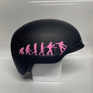 May include: Black ski helmet with a pink evolution of man sticker. The sticker shows a progression of humans from ape to snowboarder.
