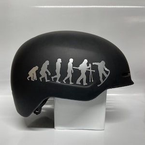 May include: A black ski helmet with a silver vinyl sticker depicting the evolution of skiing. The sticker shows a progression of stick figures, starting with a primitive ape-like figure and ending with a skier in a modern ski stance.