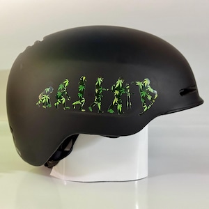 May include: A matte black helmet with a green and white graphic depicting the evolution of humans, with each stage represented by a cannabis leaf. The helmet is designed for sports or recreational activities.