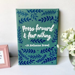 May include: A teal canvas art print with the quote "Press forward & fear nothing" in white script, framed by blue leaf designs. The quote is attributed to St. Katharine Drexel. A small pink framed picture and a vase of white flowers are in the background.