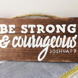 May include: A wooden sign with the text "BE STRONG & COURAGEOUS" in white paint. The text "JOSHUA 1:9" is also written in white paint below the main text.