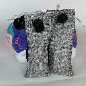 May include: Two gray fabric bags with black drawstring closures. The bags are designed to absorb odors and are placed inside a pair of colorful sneakers.
