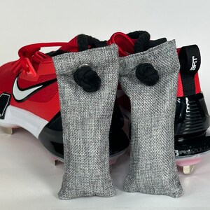 May include: Two gray fabric bags with black drawstrings, each with a metal ring. The bags are placed inside a pair of red and black baseball cleats.