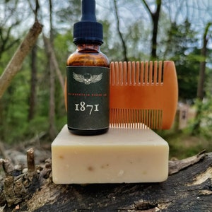 May include: A brown glass bottle with a black dropper top and a skull and wings logo. The bottle is labeled "The Mountain Beard Co. 1871". A bar of soap and a wooden comb are also in the image.