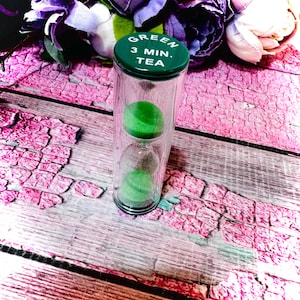 May include: A clear glass hourglass with green sand inside. The green lid is labeled "GREEN 3 MIN. TEA". The hourglass is designed to measure a 3-minute tea brewing time. The background features purple flowers and a pink wooden surface.