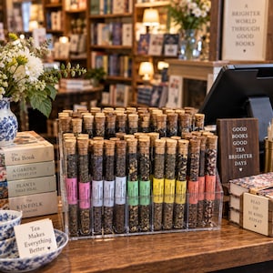 May include: A clear acrylic display stand holds multiple glass test tubes filled with various loose-leaf tea blends, each sealed with a cork stopper and labeled with a colored band. The display is surrounded by books, a floral vase, and signs with text including Tea Makes Everything Better.