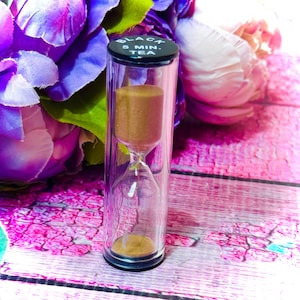 May include: A clear glass hourglass with brown sand, labeled "BLACK 5 MIN. TEA" on a black cap. The hourglass is set against a backdrop of purple and pink flowers, creating a visually appealing contrast. The timer is designed for tea brewing.