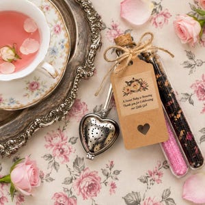 May include: A white porcelain teacup filled with pink liquid, a silver heart-shaped tea infuser, and two glass test tubes containing loose leaf tea and pink sugar. A brown paper tag with a teapot illustration and the text A Sweet Baby is Brewing Thank you for Celebrating our Little Girl is tied to the tubes with twine.