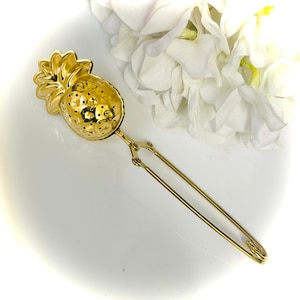 Pode incluir: Um infusor de chá dourado em forma de abacaxi, com um corpo perfurado e uma alça longa. O infusor é feito de metal e tem um design detalhado. O fundo apresenta flores brancas.