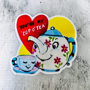 Pode incluir: Um autocolante extravagante com uma chaleira e uma chávena sorridentes. A chaleira é decorada com flores cor-de-rosa e detalhes azuis. Inclui-se também um coração vermelho com o texto "You're my Cup O' Tea".