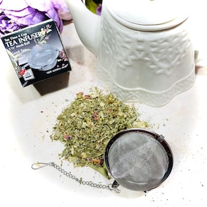 May include: A stainless steel tea infuser with a 2 1/2 inch mesh ball and a chain. The infuser is sitting on a white surface with loose leaf tea and a white teapot.