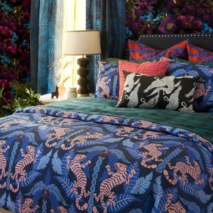 Op de afbeelding: Een slaapkamer met een bed met een blauwe en roze tijgerprint dekbedovertrek. Het bed heeft een teal groene hoofdeinde en verschillende kussens met een zwart-witte vogelprint. Een groene fluwelen stoel met een gele deken staat op de voorgrond.