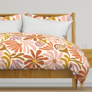 May include: A bed with a wooden frame and white pillows. The bedding features a floral pattern with large flowers in shades of orange, pink, and yellow on a cream background. The overall theme is a retro, bohemian style.