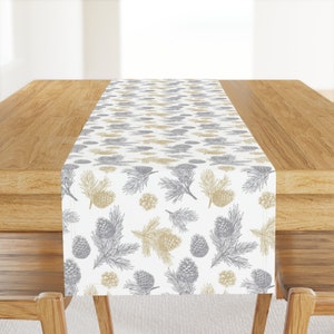 May include: A white table runner with a repeating pattern of gray and gold pine branches and pine cones.