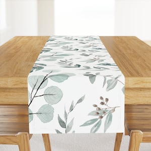 May include: A white table runner with a watercolor pattern of green leaves and branches. The runner is on a wooden table.