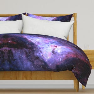 Op de afbeelding: Een dekbedovertrek en bijpassende kussenslopen met een levendige ruimte-nevelprint in tinten dieppaars, magenta en zwart, bezaaid met heldere witte sterren. De beddengoedset wordt getoond op een lichtgekleurd houten bedframe.