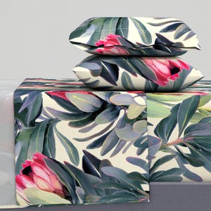 May include: Bedding set featuring a floral pattern with pink protea flowers and green leaves on a cream background. Two pillows are stacked on top of the folded sheet set. The bedding set has a botanical theme.