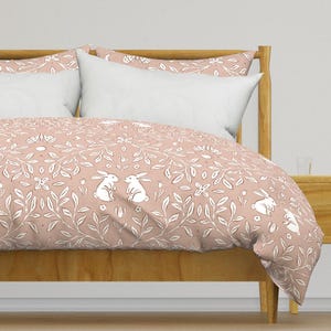 May include: A blush pink duvet cover with a white floral pattern featuring bunnies. The duvet cover is on a bed with a wooden headboard and a white pillow.