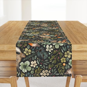 Può includere: Runner da tavolo decorativo con sfondo scuro e design ad acquerello di flora e fauna della foresta. Il design include conigli, uccelli, fiori e fogliame nei toni del verde, arancione e bianco. Il runner è su un tavolo di legno.