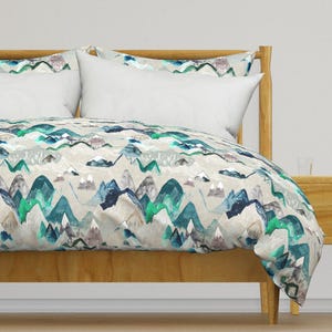 May include: A duvet cover featuring a watercolour mountain print in shades of blue, green, and grey on a beige background. The duvet cover is on a bed with a wooden frame.