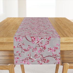 May include: A gray table runner with a pink floral pattern. The pattern features delicate branches with pink blossoms and leaves. The runner is perfect for adding a touch of elegance to any dining table.