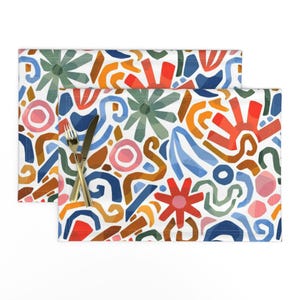Puede incluir: Un mantel individual rectangular con fondo blanco y un patrón abstracto colorido de formas rojas, naranjas, azules, verdes y rosas, que incluye flores en forma de estrella y líneas curvas, con un tenedor y un cuchillo de plata encima.