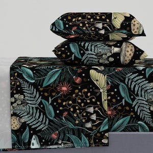 Puede incluir: Una sábana de cama negra con un patrón floral verde oscuro, dorado y rosa con setas, polillas y helechos. La sábana se muestra con fundas de almohada a juego.
