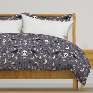 Könnte beinhalten: Dunkelgraue Bettdecke mit einem sich wiederholenden Muster aus Totenköpfen, Raben, Blitzen und floralen Elementen. Das Bettwäsche-Set enthält weiße Kissenbezüge und wird auf einem Holzrahmen präsentiert. Das Gesamtthema ist gotisch und gruselig.