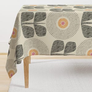以下が含まれることがあります： オレンジとピンクの中心を持つ、スタイリッシュな黒い花が繰り返しパターンで描かれたベージュのテーブルクロス。花は太陽の光線のようなデザインです。