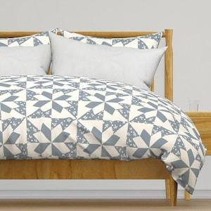 May include: A bed with a wooden frame and a duvet cover featuring a geometric pattern in shades of blue and cream. The pattern includes star-like shapes and small floral accents. Two white pillows are visible.