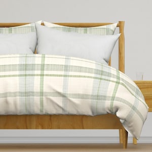 May include: A bed with a light wood frame and a duvet cover featuring a plaid pattern in shades of green and white. Two white pillows are visible, along with a wooden nightstand.