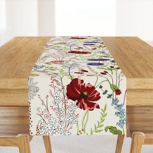 May include: A rectangular table runner featuring a cream-colored background with a botanical print of red poppies, small white flowers, blue cornflowers, and various green foliage and grasses arranged in a scattered, naturalistic pattern.