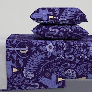Könnte beinhalten: Ein dunkelviolettes Bettwäsche-Set mit einem floralen Muster mit Schlangen, Vögeln und Monden. Das Set enthält zwei Kissenbezüge, die auf einem gefalteten Laken gestapelt sind. Das Design beinhaltet Lila-, Weiß- und Goldtöne.