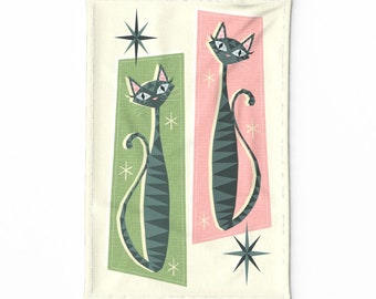 May include: A kitchen towel with a retro design featuring two stylized black cats with green and pink backgrounds. The cats have white eyes and are outlined in black. The background is a cream color with a starburst design.