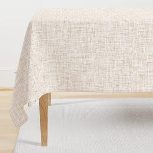 May include: A beige and white textured tablecloth draped over a wooden table. The fabric has a woven appearance, creating a neutral and versatile aesthetic. The table is set on a white rug.