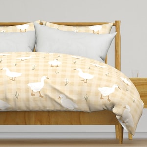 Puede incluir: Una cama con estructura de madera clara y almohadas blancas. La funda nórdica presenta un patrón repetido de gansos blancos sobre un fondo a cuadros amarillo y blanco. El tema general es de estilo granja o campestre.