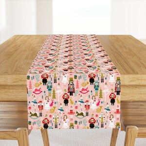 May include: A pink table runner with a repeating pattern of nutcracker characters, dancers, and Christmas trees. The pattern is in shades of red, green, blue, and gold.