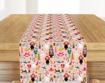 Peut inclure: Un chemin de table rose avec un motif répété de personnages de casse-noisette, de danseurs et de sapins de Noël. Le motif est dans des tons de rouge, vert, bleu et or.
