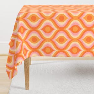 Puede incluir: Un mantel de estilo retro con un patrón repetitivo de formas anaranjadas, rosas y crema en forma de ojo. El mantel está sobre una mesa de madera, con una alfombra blanca en primer plano. El borde superior presenta un diseño geométrico.