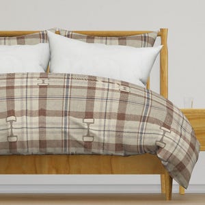 May include: A bed with a wooden frame and a plaid patterned comforter and pillowcases. The comforter features a brown, beige, and blue plaid design with decorative metal accents. White pillows rest against the headboard.