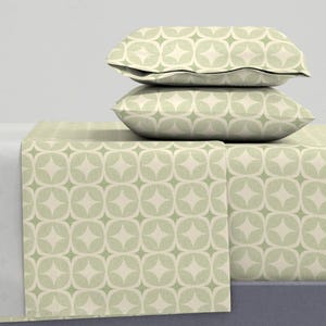 May include: A bedding set featuring a cream and light green geometric pattern. The set includes two pillowcases and a fitted sheet. The pattern consists of a series of interconnected diamond shapes within a circular design. The overall aesthetic is modern and minimalist.