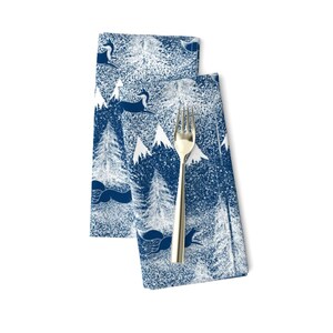Puede incluir: Dos servilletas azules con un diseño de escena de invierno blanco. El diseño presenta árboles estilizados, montañas y ciervos. Las servilletas están dobladas y un tenedor dorado se coloca en la parte superior.