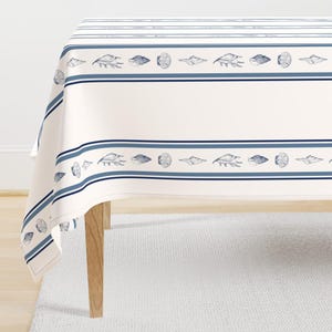 Può includere: Tovaglia color crema con un design nautico. La tovaglia presenta strisce blu e un motivo ripetuto di conchiglie. I bordi sono rifiniti con un bordo blu, creando un'estetica costiera.