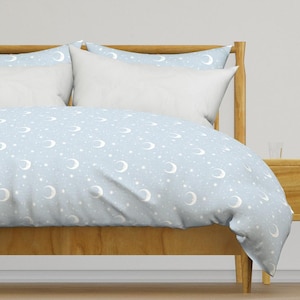 Op de afbeelding: Een lichtblauwe dekbedovertrek met een wit halve maan en ster patroon. De dekbedovertrek is op een houten bedframe met twee witte kussens.