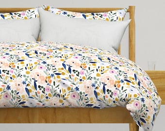 Op de afbeelding: Een witte dekbedovertrek met een bloemenpatroon met roze, gele en blauwe bloemen op een witte achtergrond. De dekbedovertrek ligt op een bed met een houten hoofdeinde en een wit kussen.