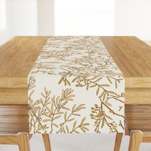 May include: A white table runner with a gold floral pattern. The pattern features delicate branches with small leaves and berries. The runner is ideal for adding a touch of elegance to any dining table.